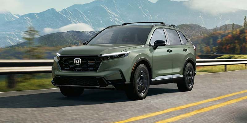 Exterior of a green 2026 Honda CR-V Hybrid SUV driving down a road with a mountain in the background near Denver, CO.
